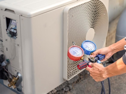 an HVAC technician servicing an outdoor air conditioning unit, specifically using a manifold gauge set to check refrigerant pressure.